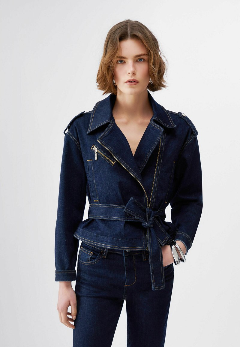 Moteris su trumpais banguotais plaukais dėvi tamsiai mėlyną džinsinę striukę su diržu juosmens srityje, užtrauktuko detalėmis, derančias džinsines kelnes ir sidabrinius apyrankes ant kairiojo riešo.