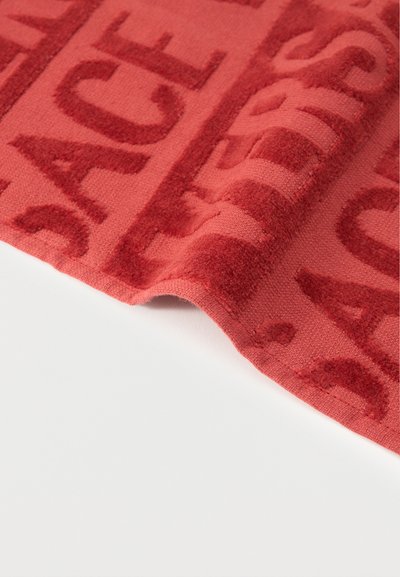 Κόκκινο υφασμάτινο με υφή, με ανεβασμένα γράμματα που σχηματίζουν μια σχεδιαστική πατρόν, μερικώς διπλωμένο πάνω σε λευκή επιφάνεια.