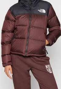 Veste matelassée bordeaux avec une partie supérieure bleu marine et une fermeture éclair. Elle présente des coutures horizontales et un logo embossé sur la poitrine.