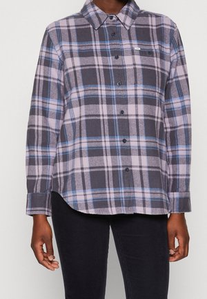 Femme portant une chemise à carreaux violette, grise et bleue à manches longues boutonnée, avec un pantalon noir, debout devant un fond blanc.