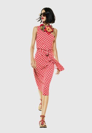 Frau geht in rotem und weiß gemustertem ärmellosem Kleid mit gebundener Taille, großer Blumenhalskette, Sonnenbrille und passenden Sandalen.