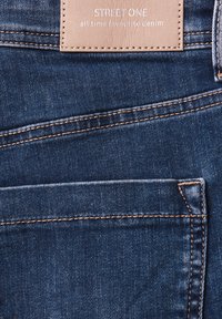 Jean en denim bleu foncé avec une surface texturée, comportant des coutures contrastées, plusieurs poches et une étiquette en cuir beige cousue à la taille.
