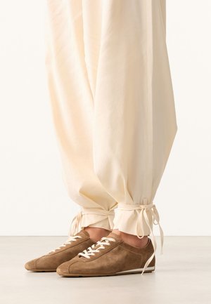 Bruine suède vetersneakers gedragen met crèmekleurige broeken, met strikjes aan de enkels, staand op een gladde lichte vloer.