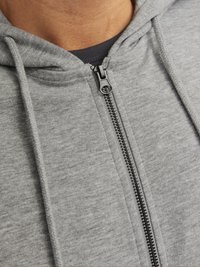 Hoodie zippé gris en matériau doux avec une finition texturée. Comprend une fermeture éclair devant et une capuche avec cordon. Superposé sur une chemise sombre.