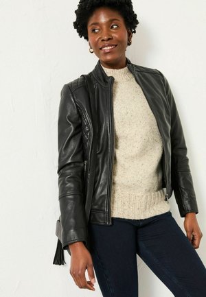 Schwarze Lederjacke mit gesteppten Schulterakzenten, getragen über einem beigen Strickpullover. Das Modell hat einen dunklen Teint und kurzes, lockiges Haar.