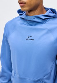 Άνδρας που φοράει ανοιχτό μπλε φούτερ Nike Pro με μαύρο λογότυπο στο κέντρο του στήθους, εμφανίζει το λαιμό και το κάτω μέρος του προσώπου.