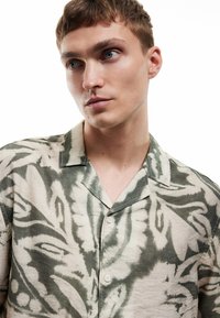 Korte mouwen shirt met een beige stof en groene bloemenpatronen, een ingesneden kraag en knoopsluiting, gemaakt van lichtgewicht materiaal.