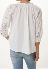 Biała bluzka z haftowanymi kwiatowymi wycięciami na rękawach i plecach, z marszczonym dekoltem i luźnym krojem. Zestawiona z jasnoniebieskimi dżinsami.