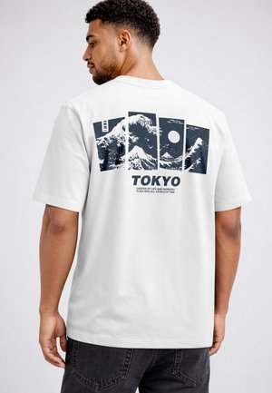 Man wearing white T-shirt with black graphic panels of waves, mountain, and moon, text reads "TOKYO CENTER OF LIFE AND HARMONY."
