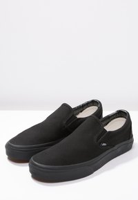 Scarpe slip-on nere con suola in gomma, caratterizzate da una tomaia in tessuto liscio e inserti elastici laterali, presentate su uno sfondo bianco.