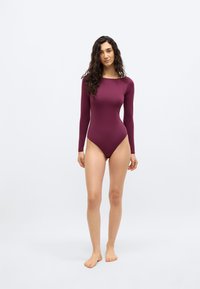 Γυναίκα με μακριά σγουρά μαλλιά, ντυμένη με εφαρμοστό μακρυμάνικο κορμάκι σε μπορντό χρώμα, όρθια, ξυπόλητη, μπροστά σε απλό, φωτεινό φόντο.