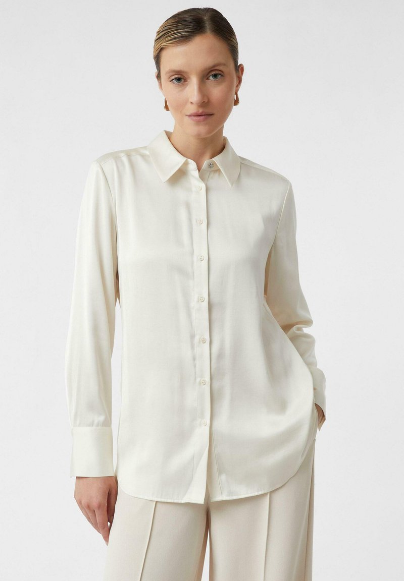 Cream-colored long-sleeve blouse with a pointed collar, front buttons, and a smooth texture. It is slightly loose-fitting with tailored sleeves.