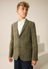 Grön tweedblazer med houndstooth-mönster, har en enkel knappstängning, två fickor och en slagkrage, tillsammans med en vit skjorta.