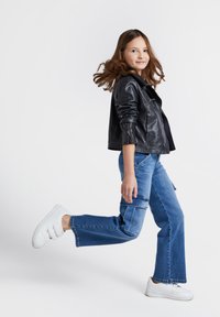 Czarna skórzana kurtka, niebieskie szerokie dżinsy z kieszeniami i białe trampki na rzepy. Osoba idzie, ma długie, falujące włosy.