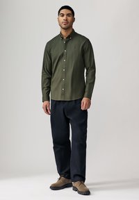 Olijfgroene langemouwen shirt met button-down kraag, witte knopen en klein rood logo, gecombineerd met donkerblauwe broek en bruine schoenen.