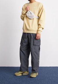 Osoba v béžové mikině Nike, šedých cargo kalhotech, olivově zelených teniskách a bílé crossbody tašce s černým logem Nike.