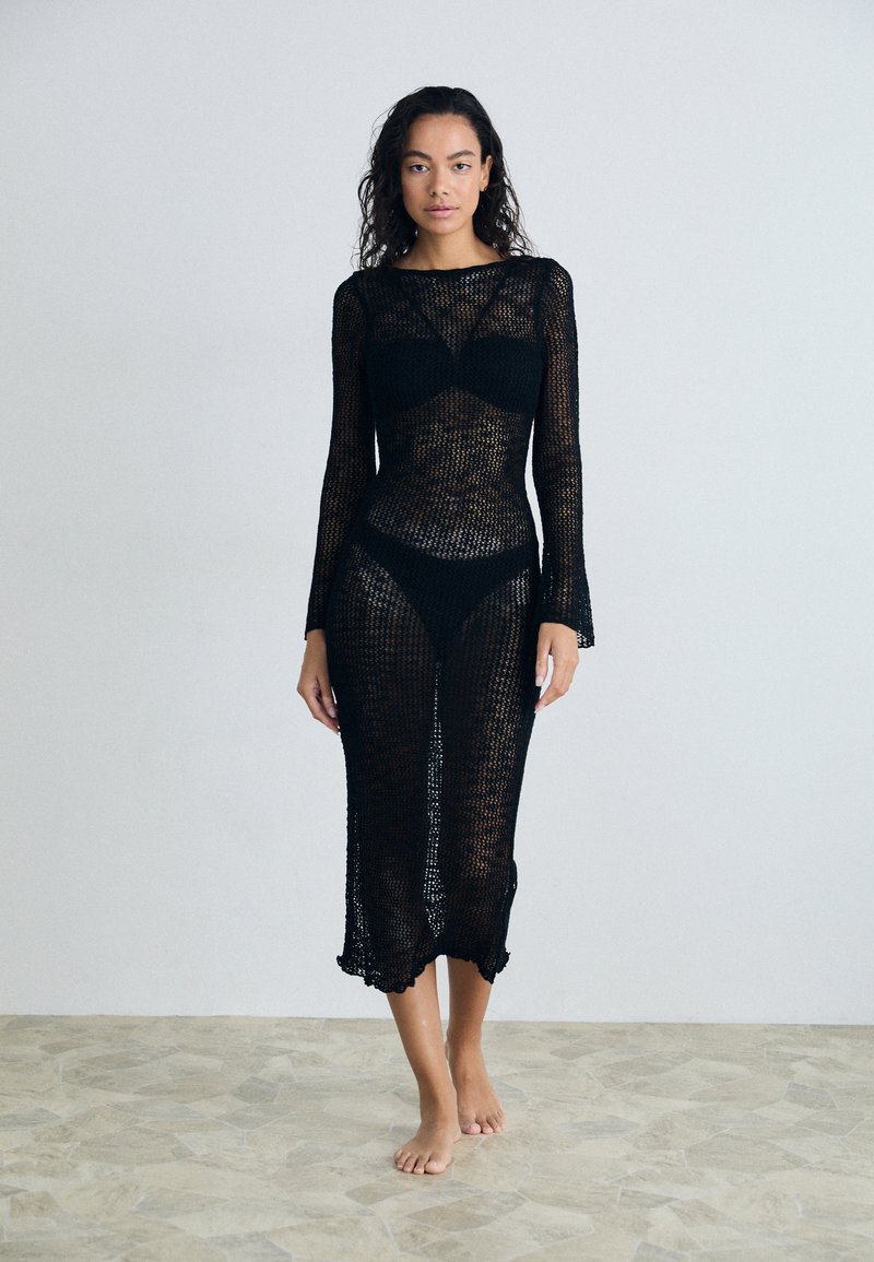Lungo abito in crochet nero con maniche lunghe e forma aderente, caratterizzato da un orlo sagomato e una texturizzazione trasparente, sovrapposto a un set bikini nero.