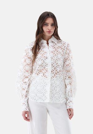 Donna con lunghi capelli castani che indossa una camicetta bianca di pizzo floreale trasparente con maniche lunghe e pantaloni bianchi su uno sfondo semplice.
