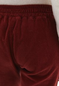 Burgundské kordové kalhoty s elastickým pasem. Texturovaná tkanina má vertikální žebra a měkký povrch. Pohled zblízka na pas a látku nohavic.