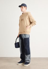 Felpa beige con cappuccio e tasca frontale, abbinata a jeans patchwork blu. Gli accessori includono una piccola borsa navy e sneakers navy.