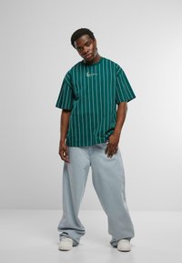 Zaļš svītrains īsām piedurknēm t-krekliņš ar apaļu kakla izgriezumu un izšūtu logo. Apvienots ar brīviem gaiši ziliem džinsiem un baltiem sporta apaviem.