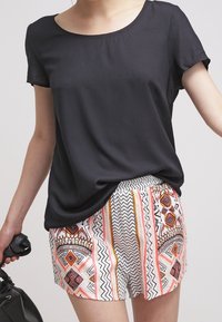 Donna che indossa un top nero a maniche corte e pantaloni corti bianchi con motivi geometrici arancioni, neri e rossi, mentre tiene una borsa nera.
