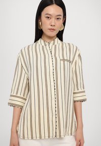 Striped shirt in cream and black, featuring a small collar, button closure, and three-quarter sleeves with trimmed accents.