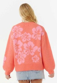 Eine Frau mit blonden Haaren trägt einen korallenfarbenen Pullover mit großen rosa Blumenmustern und einen hellblauen Jeansrock, während sie von der Kamera weg schaut.