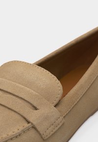 Μπεζ μοκασίνι από σουέτ με σχέδιο μοκ toe και λεπτομέρεια με λουρί στο μπροστινό μέρος, που δείχνει τις ραφές και το λείο εσωτερικό φόδρα.