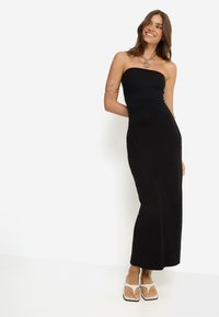 Μαύρο μάξι φόρεμα χωρίς τιράντες με εφαρμοστή σιλουέτα. Λείο υλικό με μακρύ τελείωμα. Συνδυάζεται με λευκά σανδάλια. Μινιμαλιστικός σχεδιασμός.