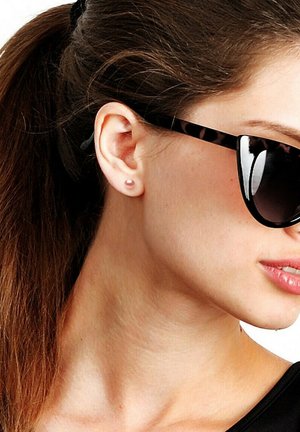 Femme aux longs cheveux bruns attachés en queue de cheval, porte des lunettes de soleil noires et de petites boucles d'oreilles roses en forme de clous, tournée vers la droite avec une expression neutre.