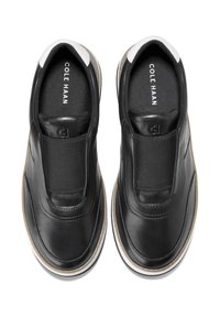 Schwarze Slipper-Sneaker mit weißen Sohlen und braunen Akzenten, elastische Bänder über dem Obermaterial, "Cole Haan" im Inneren jeder Ferse bedruckt.