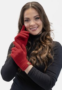 Røde strikkede handsker med ribbede manchetter, der har et lille brunt mærke. Handskerne bæres af en model i en sort turtleneck mod en hvid baggrund.