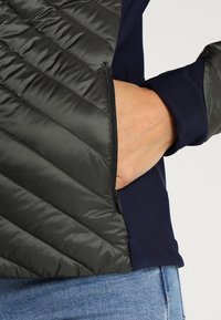 Kvadrofilt mörkgrön puffväst med en marinblå långärmad skjorta under, som har en sidficka med dragkedja.