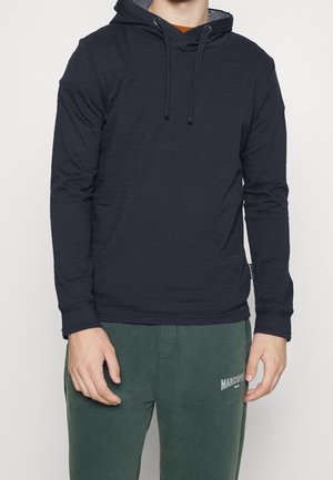 Sudadera con capucha de un azul marino oscuro, con mangas largas, tela texturizada y cordones. Combinada con pantalones de chándal verdes que presentan un logo.