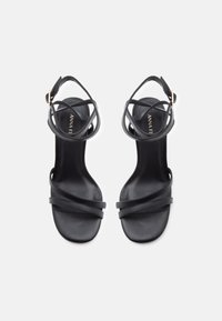 Musta värvi kõrge kontsaga sandaalid, ristuvad rihmad ja pahkluu pannal. Sile tekstuur, avatud varvaste disain ja nähtav brändiga sisetald.