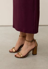 Aranyszínű magassarkú szandálok texturált felsőrésszel, bokapánttal és blokksarokkal. Egy sötét bordó ruhával viselve.