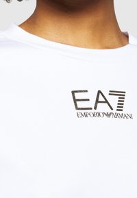 T-shirt in cotone bianco con logo nero riportante il testo "EA7" e "Emporio Armani". Collo rotondo e morbido al tatto.