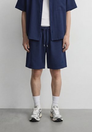 Person som har på seg marineblå shorts med snor, matchende kortermet skjorte, hvite sokker og hvite joggesko, står mot en ensfarget vegg.