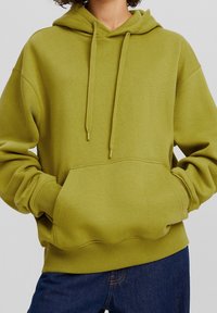 Persona con una sudadera verde oliva con bolsillo frontal y cordones, manos en los bolsillos, combinada con jeans de mezclilla azules.