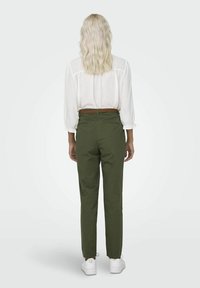 Pantaloni verde oliva con un design aderente, passanti per cintura e una cintura marrone texturizzata. Indossati con una camicia bianca a maniche lunghe e sneakers bianche.