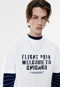 Witte t-shirt met korte mouwen en zwarte verouderde tekst "FLIGHT PB19 WELCOME TO CHICAGO," gedragen over een marineblauwe gestreepte lange mouwen shirt.