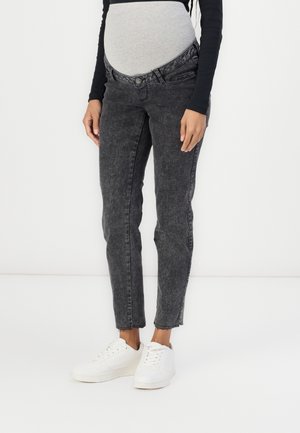 Personne enceinte portant un haut noir à manches longues, un jean de maternité gris avec un panneau élastique au niveau du ventre, et des baskets blanches, debout devant un fond blanc.