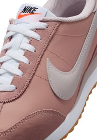 Zapatilla Nike en rosa claro y gris, con materiales de ante y tela, cordones blancos y una suela de goma con un patrón texturizado.