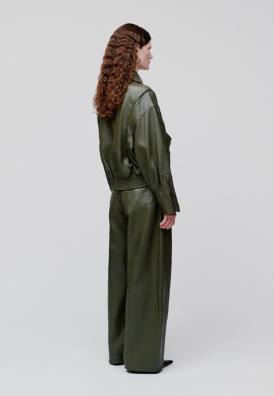 Femeie cu păr lung și creț, purtând o jachetă verde de piele și pantaloni evazați, stând pe partea laterală, pe un fundal simplu.