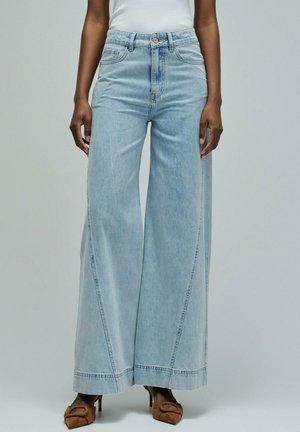 Femme portant un jean taille haute à jambes larges de couleur bleu clair, associé à des chaussures à talons pointues marron et un haut blanc sans manches.