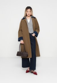 Young woman in brown coat with navy cuffs, gray zip-up sweater, dark wide-leg jeans, red flats, holding a brown handbag, standing against white background.