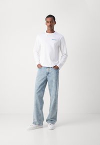 Tricou alb cu mânecă lungă, având un mic logo bleu, asortat cu blugi de denim albaștri deschis și adidași albi. Croi casual, siluetă relaxată.