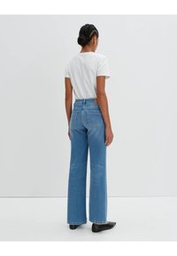 Kvinna med flätat hår iförd en vit t-shirt, högmidjade blå jeans och svarta platta skor, stående mot en enfärgad ljus bakgrund.