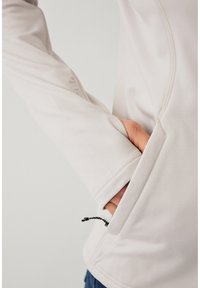 Mano deslizándose en el bolsillo delantero de una chaqueta gris claro, mostrando detalles de costura en la manga y el bolsillo con un tirador de cremallera negro visible.
